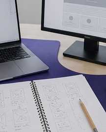 Desk with a code editor, a monitor showing a wireframe and a notebook of page sketches